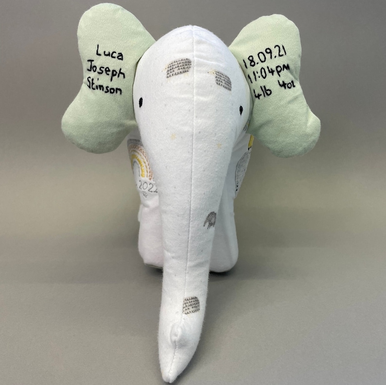 May include: A white stuffed elephant with green ears. The elephant has a rainbow design on its body and the text "Luca Joseph Stinson 18.09.21 11:04pm 4lb 4oz" is embroidered on the ears.