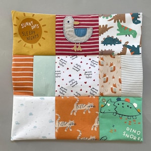 May include: A square patchwork quilt featuring nine distinct fabric panels. Patterns include a yellow sun with text reading Sunny Days Sleepy Nights, a seagull, dinosaurs, foxes, tigers, and text reading daddy + mummy love me with hearts.