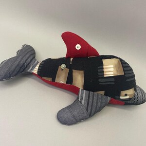 May include: A handmade stuffed dolphin toy with a red fin and black, gray, and brown fabric patchwork. The dolphin has a button on its back.