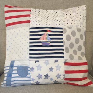 May include: A patchwork cushion with a nautical theme. The cushion is made from different fabrics with various patterns, including stripes, stars, and polka dots. A small sailboat is embroidered in the centre of the cushion.