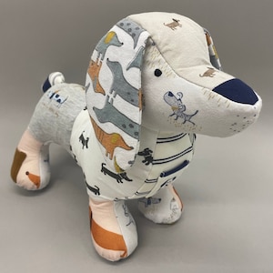 May include: A stuffed dachshund dog toy made from various patterned fabrics. The dog features a white body with black, orange, and gray dachshund illustrations. The ears and head are a mix of beige and patterned fabrics. The nose is navy blue.