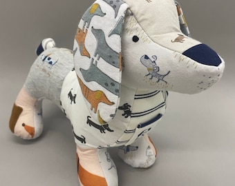 May include: A stuffed dachshund dog toy made from various patterned fabrics. The dog features a white body with black, orange, and gray dachshund illustrations. The ears and head are a mix of beige and patterned fabrics. The nose is navy blue.