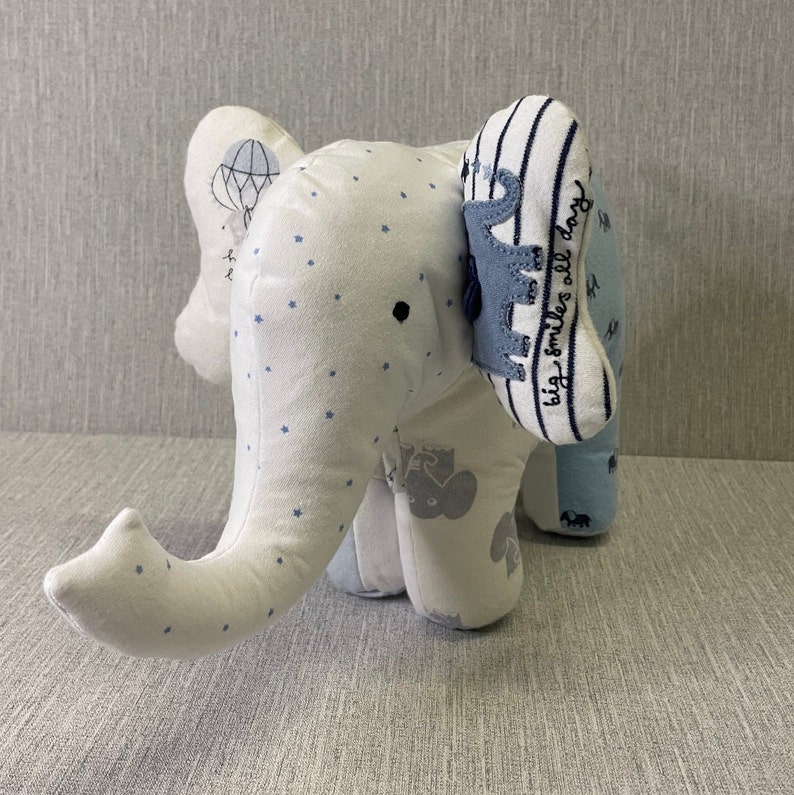 May include: A white stuffed elephant toy with blue star print fabric. The elephant has a blue and white striped ear with the text "big smiles all day".