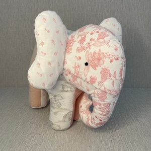 May include: A pink and white fabric elephant stuffed animal with a floral pattern. The elephant has a small black button for an eye.