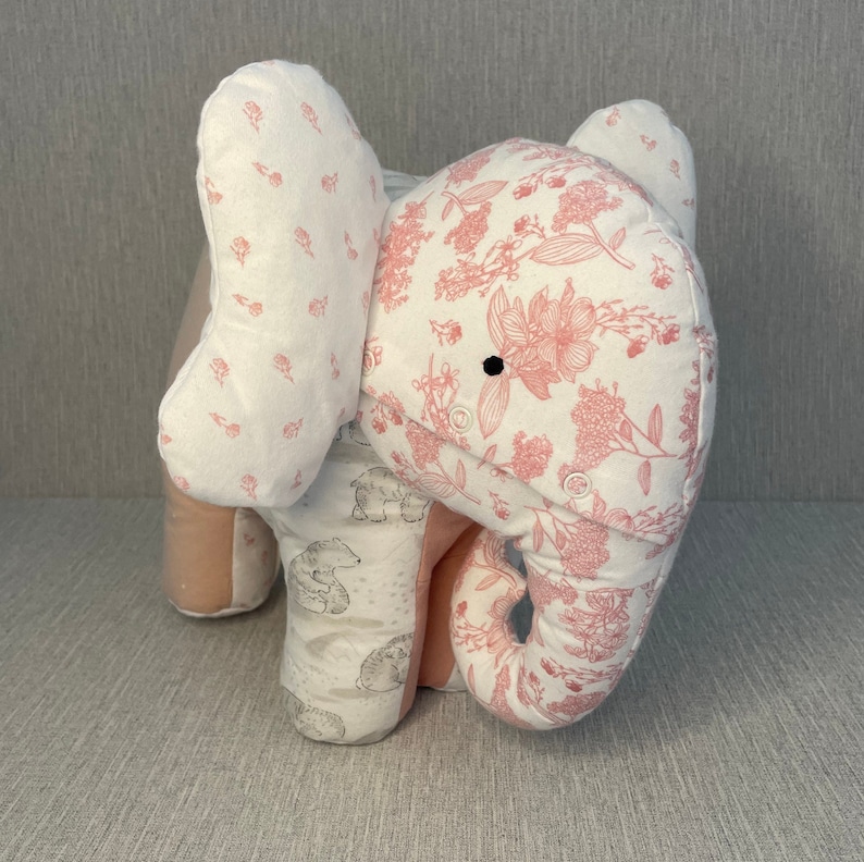 May include: A pink and white fabric elephant stuffed animal with a floral pattern. The elephant has a small black button for an eye.