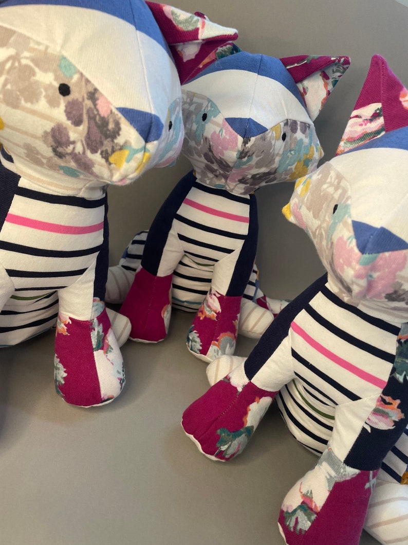 May include: Three plush toys shaped like foxes, each with a different floral pattern on their bodies. The foxes are made of a patchwork of fabrics, including blue and white striped fabric, pink and white striped fabric, and floral fabric. The foxes are sitting on a gray surface.