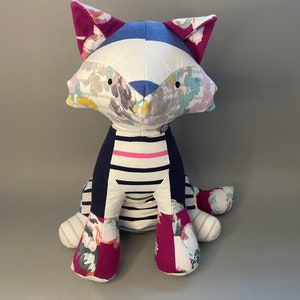 May include: A handmade plush toy cat with a white, blue, and pink floral patterned fabric. The cat is sitting with its legs crossed and has a striped fabric on its torso.