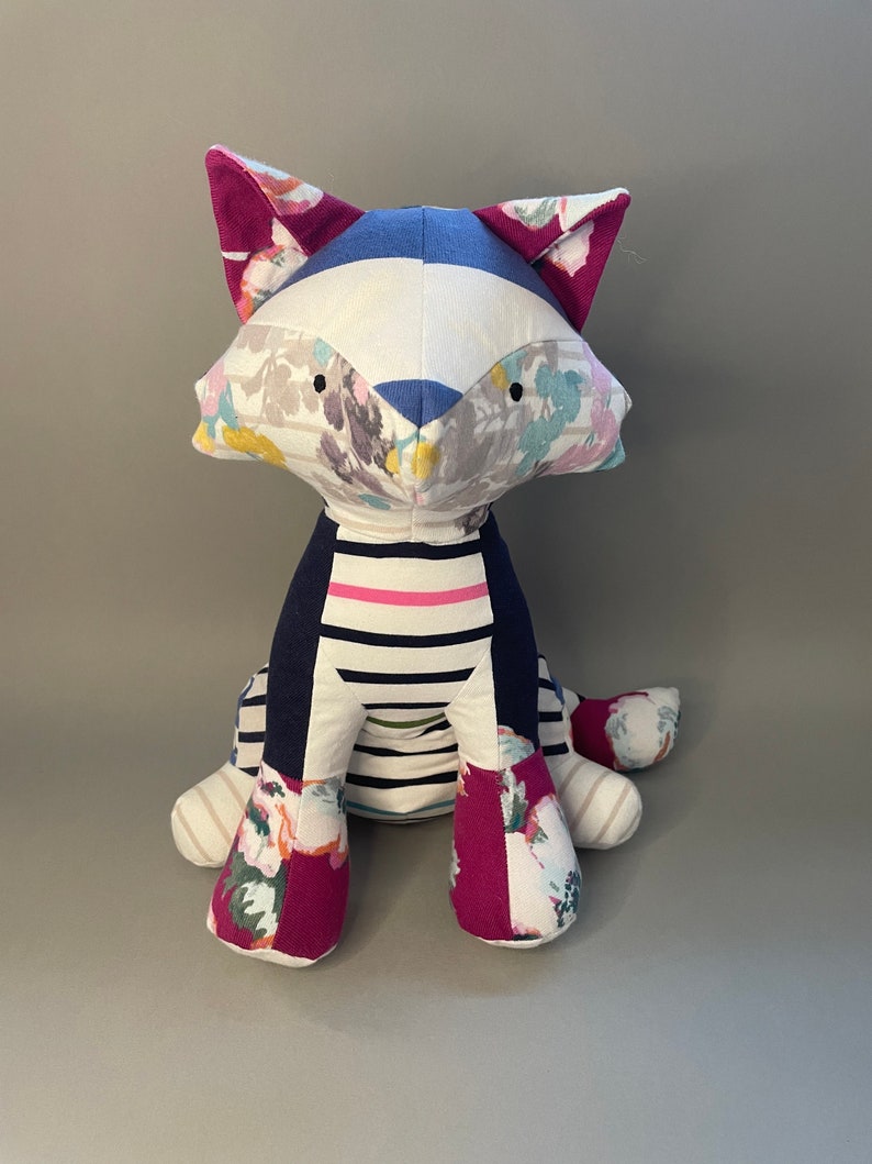 May include: A handmade plush toy cat with a white, blue, and pink floral patterned fabric. The cat is sitting with its legs crossed and has a striped fabric on its torso.