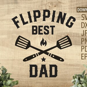 May include: Black and white graphic design with the text "Flipping Best Dad" and a pair of crossed spatulas with a flame and star in the center.