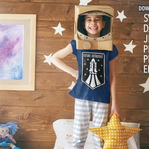 May include: A child wearing a blue t-shirt with a white rocket ship graphic and the text "Future Astronaut". The child is wearing a cardboard astronaut helmet and holding a yellow star-shaped pillow.