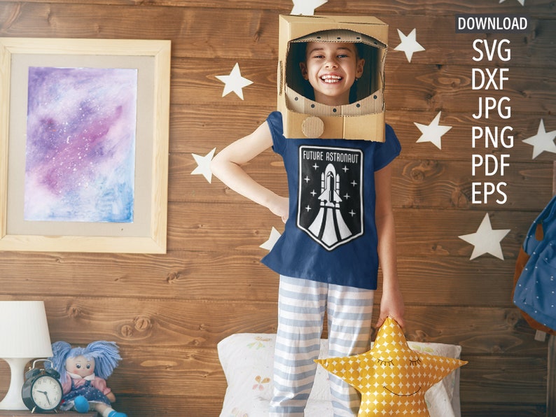 May include: A child wearing a blue t-shirt with a white rocket ship graphic and the text "Future Astronaut". The child is wearing a cardboard astronaut helmet and holding a yellow star-shaped pillow.