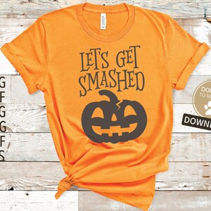 Puede incluir: Camiseta naranja con el texto "Let's Get Smashed" y una silueta negra de una calabaza rota. La camiseta está sobre un fondo de madera.