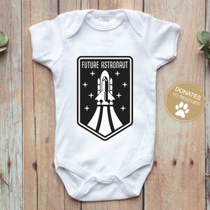 May include: White baby onesie with a black and white graphic of a rocket ship and the text "FUTURE ASTRONAUT". A small paw print is in the lower right corner with the text "DONATES TO RESCUES".