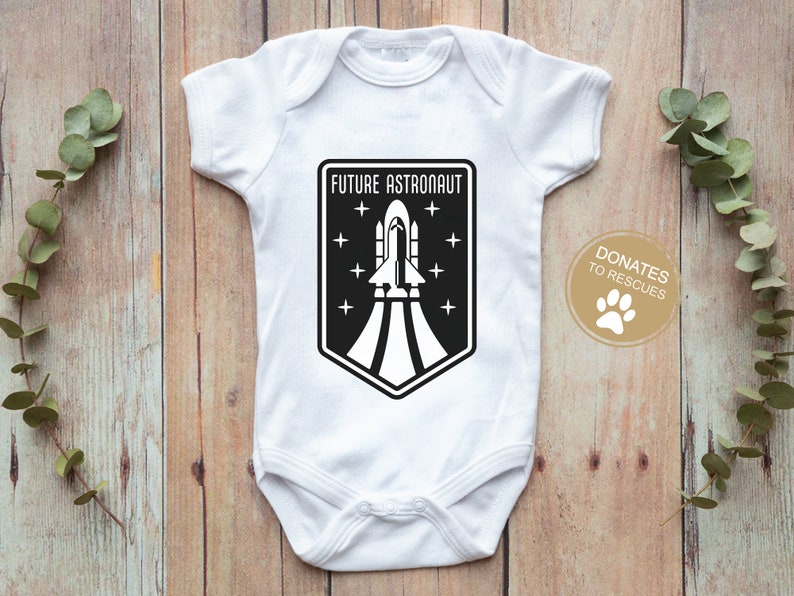 May include: White baby onesie with a black and white graphic of a rocket ship and the text "FUTURE ASTRONAUT". A small paw print is in the lower right corner with the text "DONATES TO RESCUES".