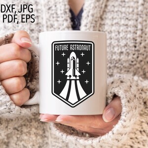 May include: White ceramic mug with a black and white graphic of a rocket ship taking off. The graphic is surrounded by a black shield with the text "FUTURE ASTRONAUT" above the rocket ship and stars around the shield.