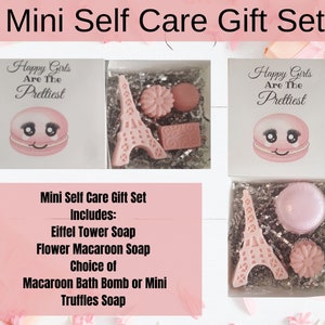 May include: Mini self-care gift set with pink soaps. The set includes an Eiffel Tower soap, flower macaroon soap, and a macaroon bath bomb or mini truffles soap. The box lid features a smiling macaroon and the text "Happy Girls Are The Prettiest."
