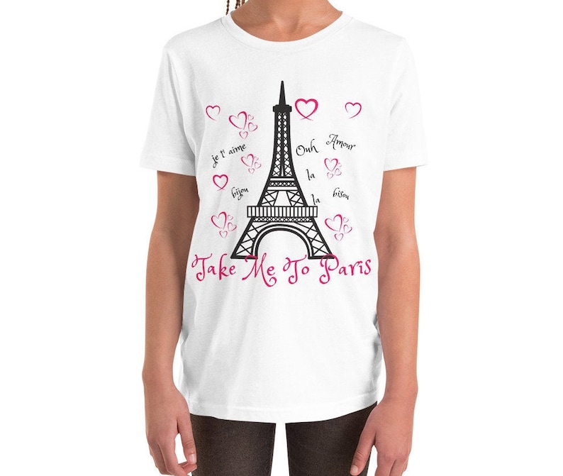 K&ouml;nnte beinhalten: Wei&szlig;es T-Shirt f&uuml;r Jugendliche mit einem schwarzen Motiv des Eiffelturms und rosa Herzen. Der Text "Take Me To Paris" ist in rosa Kursivschrift unter dem Turm geschrieben. Das Motiv enth&auml;lt auch franz&ouml;sische S&auml;tze wie "Je t'aime" und "Bisous".