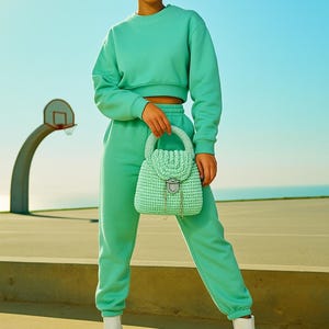 May include: A person wearing a mint green cropped sweatshirt and matching sweatpants, holding a mint green crocheted handbag. The person is also wearing white heeled boots. The background is a basketball court and a clear blue sky.