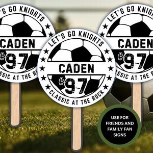 May include: Three white and black round signs with a soccer ball graphic, the text "Let's Go Knights", a name, and a number. The signs are on wooden sticks and are on a green grassy surface with a soccer ball in the background. The text on the signs reads "Let's Go Knights", "Caden #97", and "Classic at the Rock".