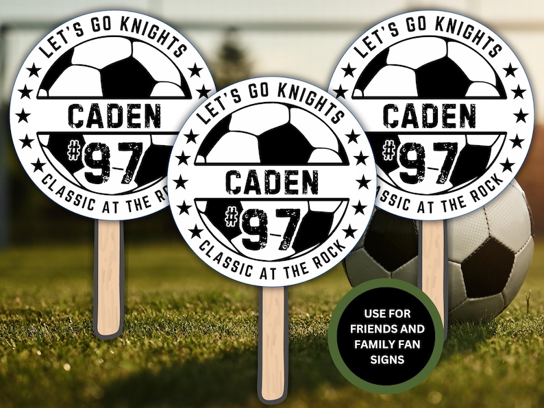 May include: Three white and black round signs with a soccer ball graphic, the text "Let's Go Knights", a name, and a number. The signs are on wooden sticks and are on a green grassy surface with a soccer ball in the background. The text on the signs reads "Let's Go Knights", "Caden #97", and "Classic at the Rock".