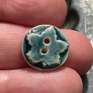 May include: A round ceramic button featuring a raised, embossed leaf design in shades of deep forest green and pale seafoam green. The button has a smooth, glossy glaze finish and includes two centered holes for attachment to fabric or garments.