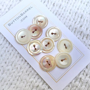 Op de afbeelding: Een set van tien witte parelmoer knopen met elk twee gaten. De knopen zijn gerangschikt op een witte kaart met de tekst "BUTTONBARREL.COM" erop gedrukt.