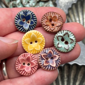Peut inclure: Six boutons ronds en céramique présentant un motif de fleur à cinq pétales en relief au centre. Les boutons sont émaillés dans diverses couleurs, dont bleu foncé, marron, jaune, vert clair, rose et un mélange violet-marron, avec deux trous.