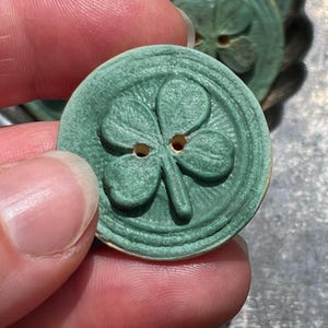 May include: A round, sage green button featuring a raised four-leaf clover design in the center. The button has two small holes for attachment and a textured, circular border surrounding the clover motif, with a matte finish on the surface.