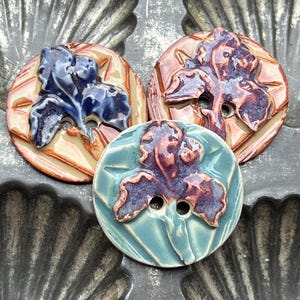 May include: Three round ceramic buttons, each featuring a raised, molded iris flower design. One button is blue and tan, one is brown and tan, and one is purple and light blue. Each button has two central holes for attachment and a glossy finish.
