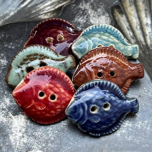 May include: A collection of six ceramic buttons shaped like fish, each featuring a glossy glaze in distinct colors including deep red, light blue, dark blue, brown, and teal. Each button has two central holes for attachment and textured scales.