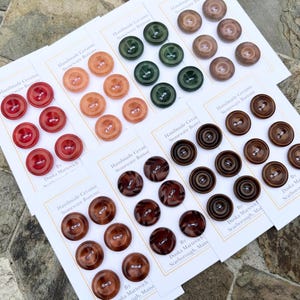 Puede incluir: Surtido de botones de cerámica artesanal de gres en varios colores y diseños, incluyendo verde, marrón y naranja. Los botones se muestran en tarjetas blancas con el texto "Botones de cerámica artesanal de gres" y el nombre del fabricante.