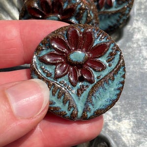 May include: A round ceramic button featuring a glossy dark red flower design centered on a textured light blue background. The button has two holes in the middle and a speckled brown border that frames the floral motif and leaf patterns.