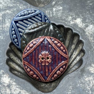 May include: Three circular ceramic buttons with a glossy finish. The top button is blue, the middle is dark gray, and the bottom is a reddish-purple with a central flower motif. Each button features a raised diamond pattern and beaded edges.