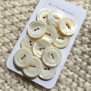 May include: A collection of round, off-white mother-of-pearl buttons with two holes, arranged on a white rectangular card labelled BUTTONBARREL. Each button features a smooth, iridescent surface and a slightly raised centre rim, showing natural variations.