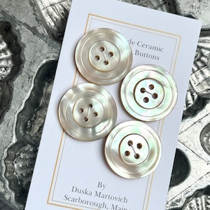 May include: Four white ceramic buttons with four holes each. The buttons have a slightly iridescent finish and are arranged on a white card with the text "de Ceramic Buttons" and "By Duska Martovich Scarborough, Maine".