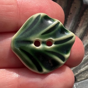 May include: A ceramic button with a glossy finish, featuring a leaf-like shape and a two-tone colour scheme of deep forest green and creamy off-white. The button has two central holes for attachment and a smooth, slightly irregular organic edge.