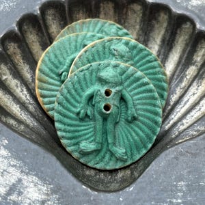 May include: Three round teal buttons featuring a raised frog figure in the center. Each button has a textured, sunburst-like pattern radiating from the frog to the edge. The buttons have two small holes in the center for attachment purposes.