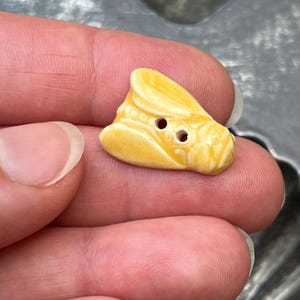 May include: A small, yellow, ceramic button shaped like a cicada or insect. The button features a glossy, smooth finish with two central holes for attachment. The insect design includes molded wing details and a segmented body structure in bright yellow.