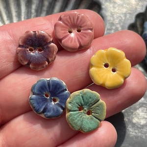 Puede incluir: Cinco botones pequeños, redondos y con forma de flor, cada uno con dos agujeros centrales. Los botones tienen un acabado esmaltado brillante en colores distintos: rosa empolvado, morado oscuro, amarillo brillante, azul oscuro y verde salvia.