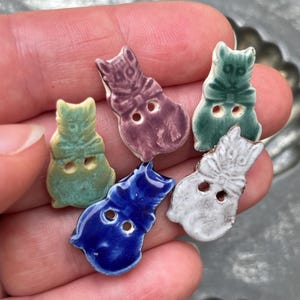 May include: Five small ceramic buttons shaped like cats, each featuring two holes for attachment. The buttons are glazed in distinct colors including light green, purple, dark green, deep blue, and white with brown edges, all showing a glossy finish.