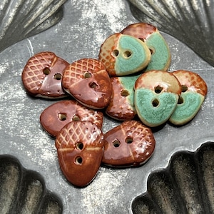 May include: A collection of ceramic buttons shaped like acorns, featuring a glossy brown finish on the textured cap and a smooth, matte teal or brown glaze on the base. Each button has two central holes for attachment, with varied color patterns.