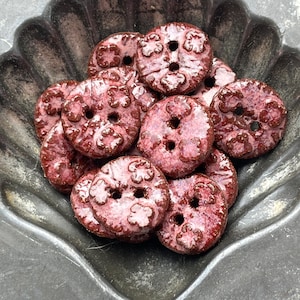 May include: A collection of round, two-hole buttons with a glossy, mottled burgundy and pink glaze. Each button features a raised, embossed floral pattern across the surface. The buttons are clustered together, showing a smooth, reflective finish.