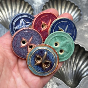 May include: A collection of six round ceramic buttons in various colors including red, green, and shades of blue. Each button features a textured surface with a raised star or cross design and two central holes for attachment to fabric or garments.
