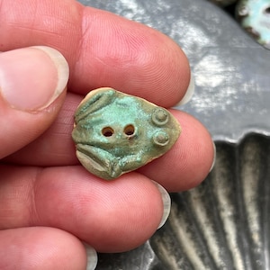 May include: A small, frog-shaped button with a weathered, verdigris-inspired green and tan finish. The button features two central holes for attachment, raised circular eyes, and molded leg details on a textured, matte surface.