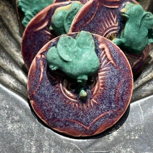May include: A set of round ceramic buttons featuring a deep purple glaze with a textured finish and a raised, matte green frog figure attached to the center. Each button has two visible holes for sewing, with a sunburst pattern etched into the surface.