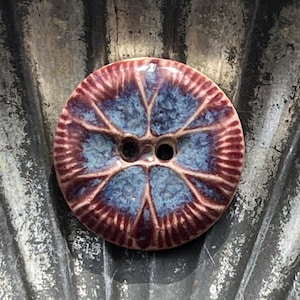 May include: A round, two-hole button featuring a glossy glaze with a central blue, textured pattern radiating outward into deep maroon, ribbed edges. The surface displays a high-shine finish with distinct color transitions between the blue center and the dark red border.