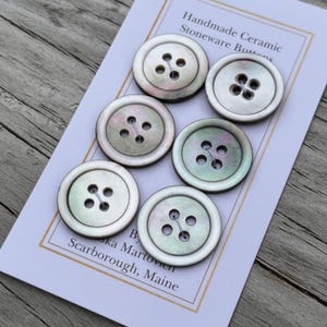 May include: Six round, four-hole buttons with an iridescent, pearlescent finish in shades of white, cream, and soft green. The buttons are arranged on a white display card featuring the text Handmade Ceramic Stoneware Buttons by Aska Martovien.