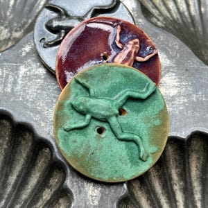 May include: Three circular ceramic buttons, each featuring a raised frog relief. One button is a matte green, one is a glossy deep purple, and one is a dark metallic gray. Each button has two central holes for attachment, showcasing a textured finish.