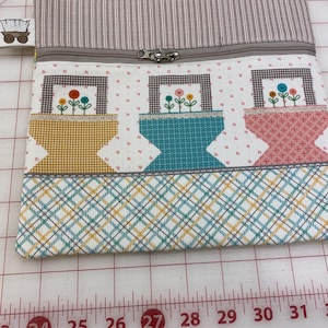 May include: A handmade zippered pouch with a floral design. The pouch features three square panels with flower pot designs in yellow, teal, and pink. The top is striped, and the bottom has a plaid pattern. A small wagon tag is attached.
