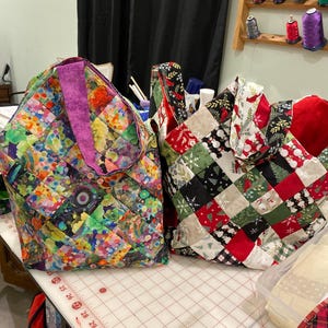 May include: Two quilted tote bags. One bag features a colorful patchwork design with a purple handle. The other bag has a patchwork pattern with red, green, and white Christmas-themed squares. Both bags are on a white surface.